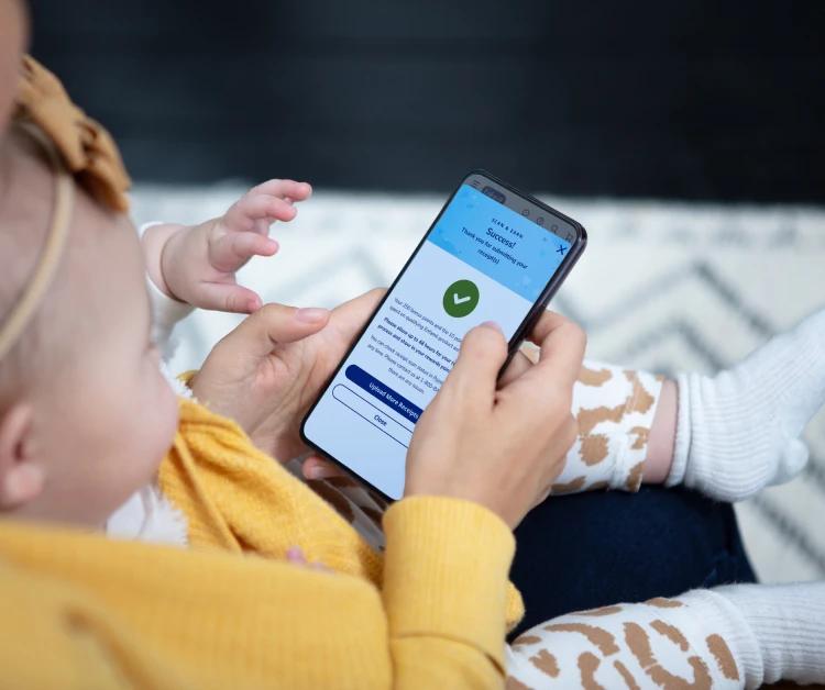 Mom holding baby and phone with phone screen displaying Enfamil app